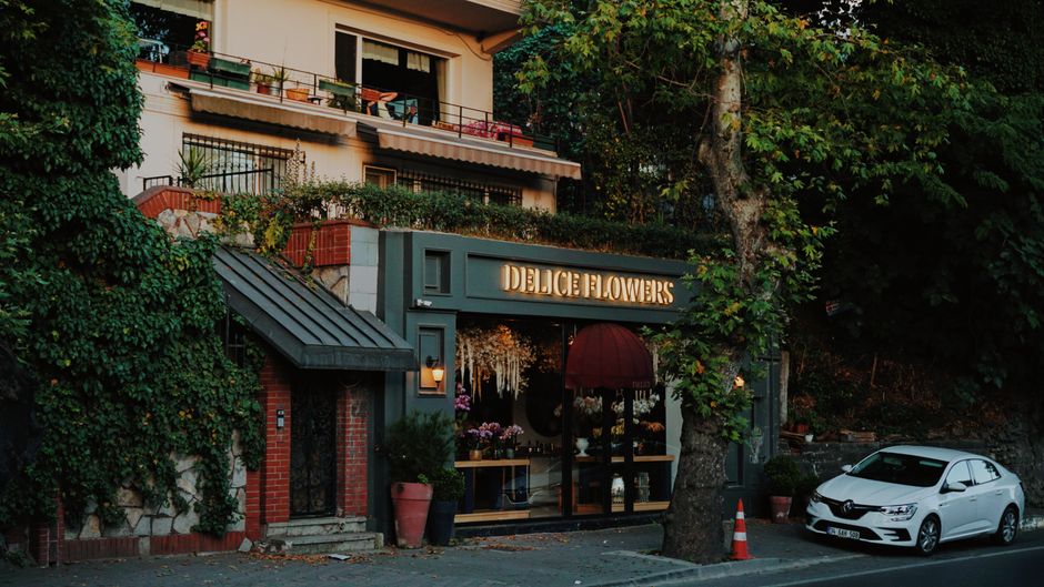 The exterior of Delice Flowers, a florist shop located on a street with lush green trees and residential buildings. The shop features a dark green façade with a prominent illuminated sign displaying