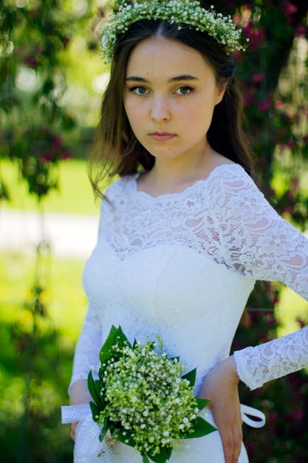A young woman with fair skin, brown hair, and blue eyes wears a white lace wedding dress with long sleeves and a floral crown made of small white flowers and greenery. She holds a bouquet of miniature
