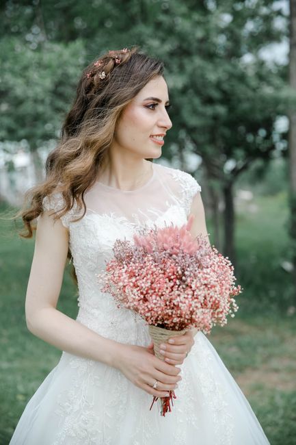 A bride with long, wavy brown hair styled with small floral accessories, wearing a delicate lace wedding dress with cap sleeves, stands outdoors in a park-like setting with lush green trees and soft n