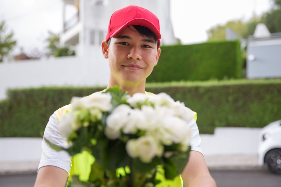 A delivery person wearing a red cap and high-visibility vest holding a bouquet of white flowers towards the camera. The bouquet features numerous white blooms with green foliage, arranged in a loose,