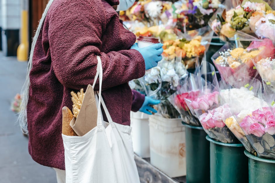 A person wearing a maroon fleece jacket and blue gloves is standing at an outdoor flower stall, holding a takeaway coffee cup in one hand and a white reusable shopping bag with a brown paper bag conta